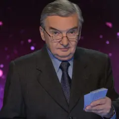 Tadeusz Sznuk w garniturze i okularach trzyma w ręku niebieskie karty na tle fioletowej, rozmytej scenografii.