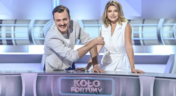 Uśmiechnięci prowadzący Koła Fortuny, mężczyzna w jasnej marynarce i kobieta w białej sukience, przy panelu z logo programu.